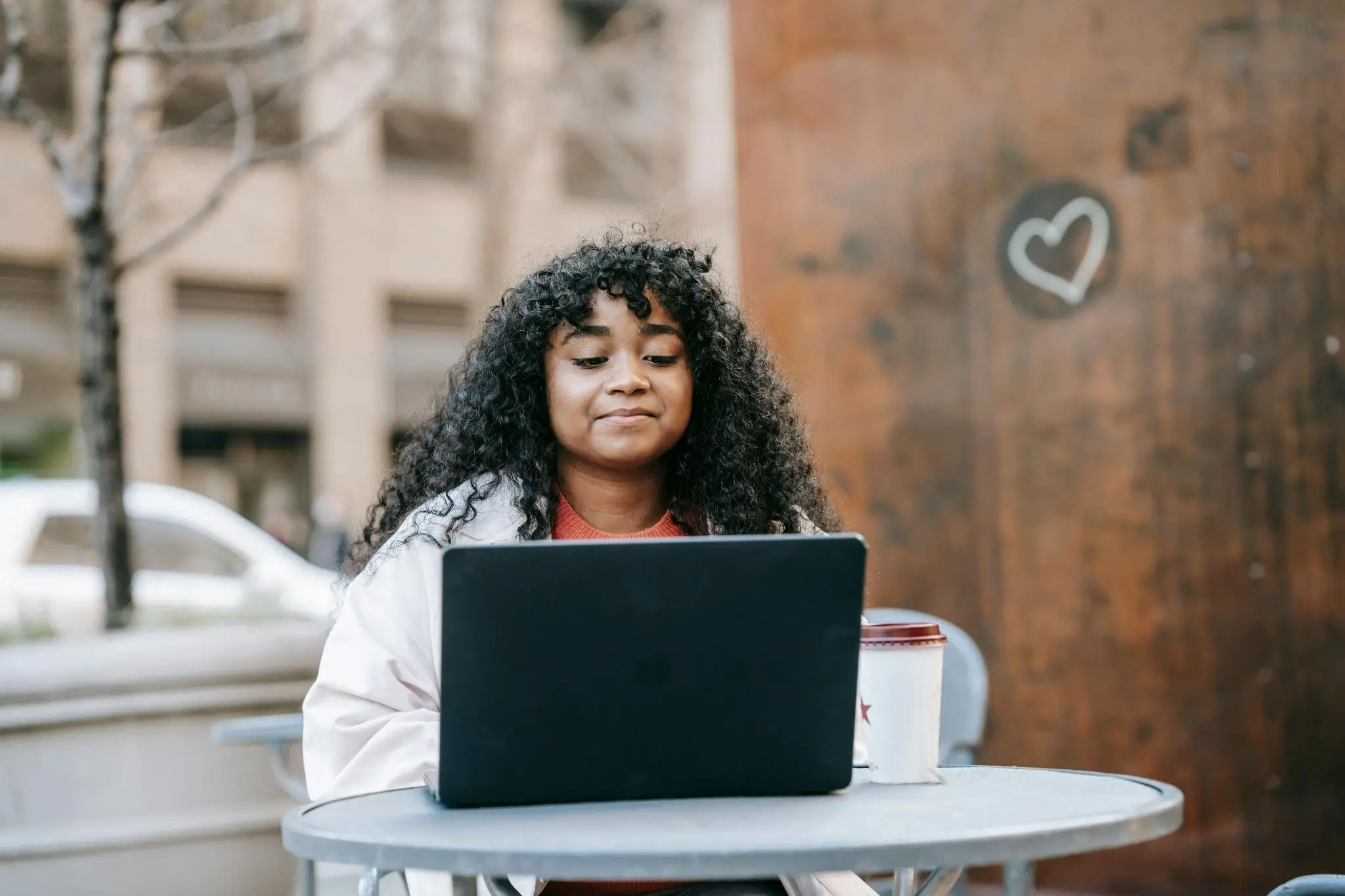 Person with curly hair using a laptop at outdoor cafe table with coffee cup