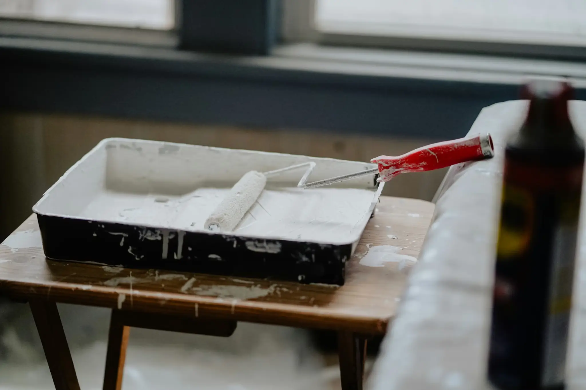 Paint roller with red handle resting in tray of white paint on wooden table