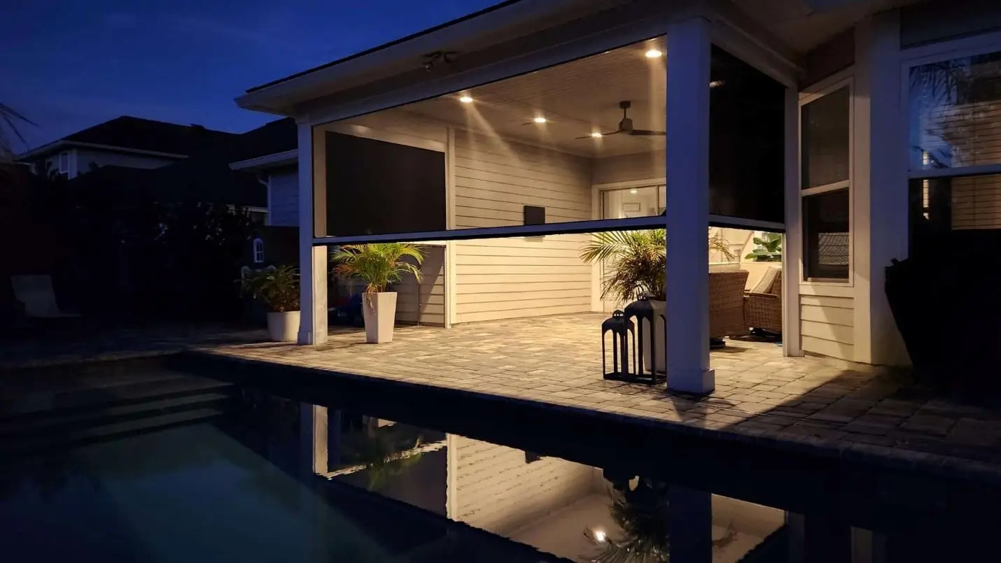 Covered patio with wicker furniture and plants, warmly lit at night by a pool