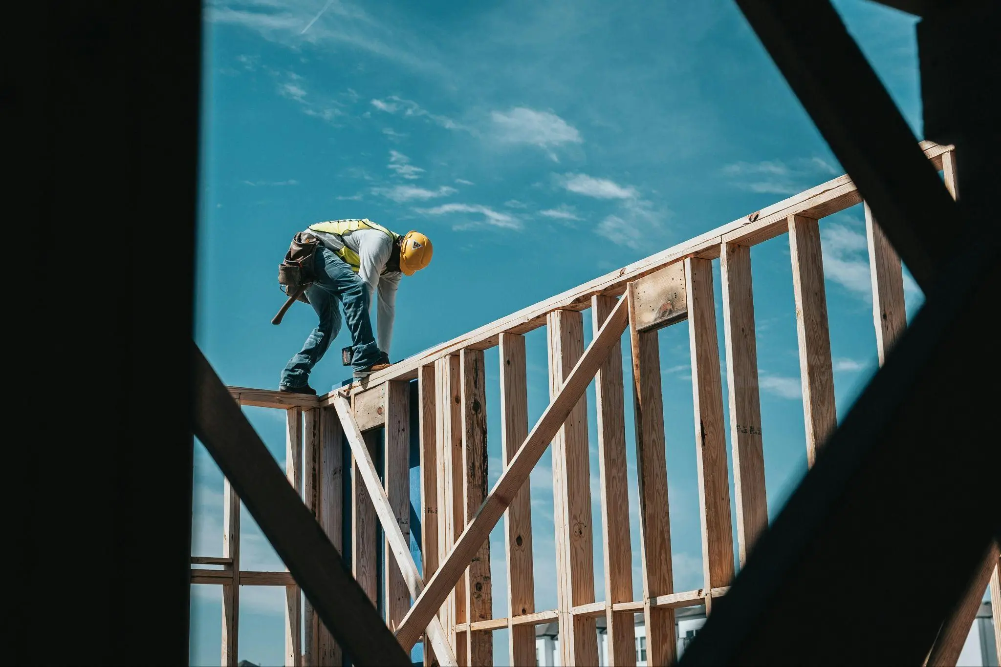 Construction worker in yellow hard hat building wooden framework under clear blue sky