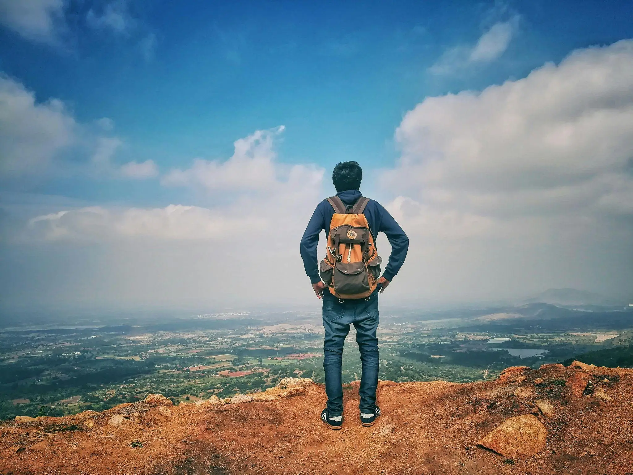Person with backpack standing on mountain summit overlooking expansive landscape under blue sky