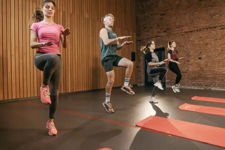 Four people exercising in a gym with wood and brick walls, performing knee lifts
