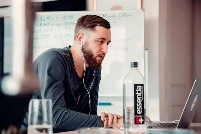 Man with earbuds using laptop at desk in modern office with whiteboard nearby