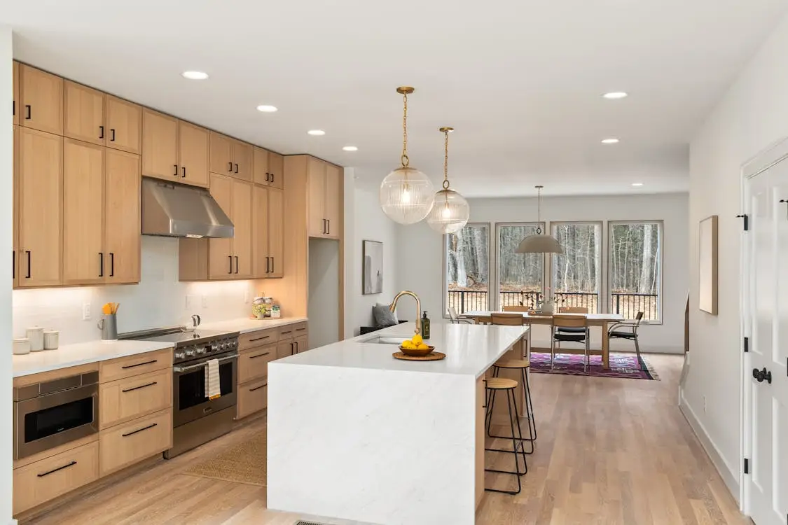 Modern kitchen with wooden cabinets, marble island, pendant lights, and dining area with large windows
