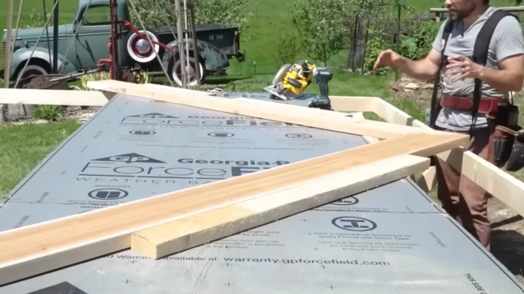 A person in a tool belt stands beside a large sheet of weather barrier material, arranging wooden planks on top