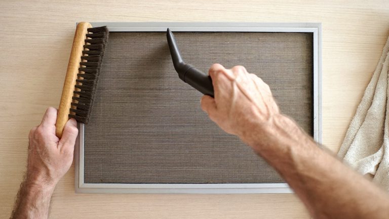 Hands cleaning a dusty HVAC air filter with brush and vacuum attachment on wooden surface.
