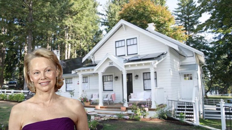 Pamela Anderson smiling in a strapless purple dress standing in front of a charming white cottage-style house surrounded by trees
