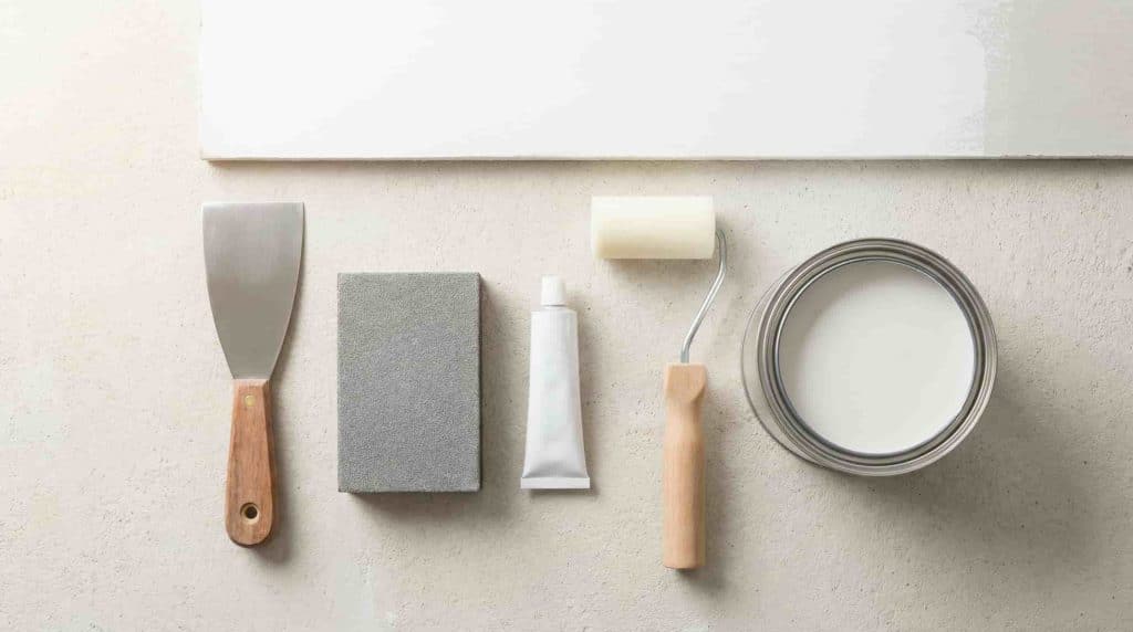 Tools for repairing chipped paint, including a putty knife, sanding sponge, filler, roller, and paint can.