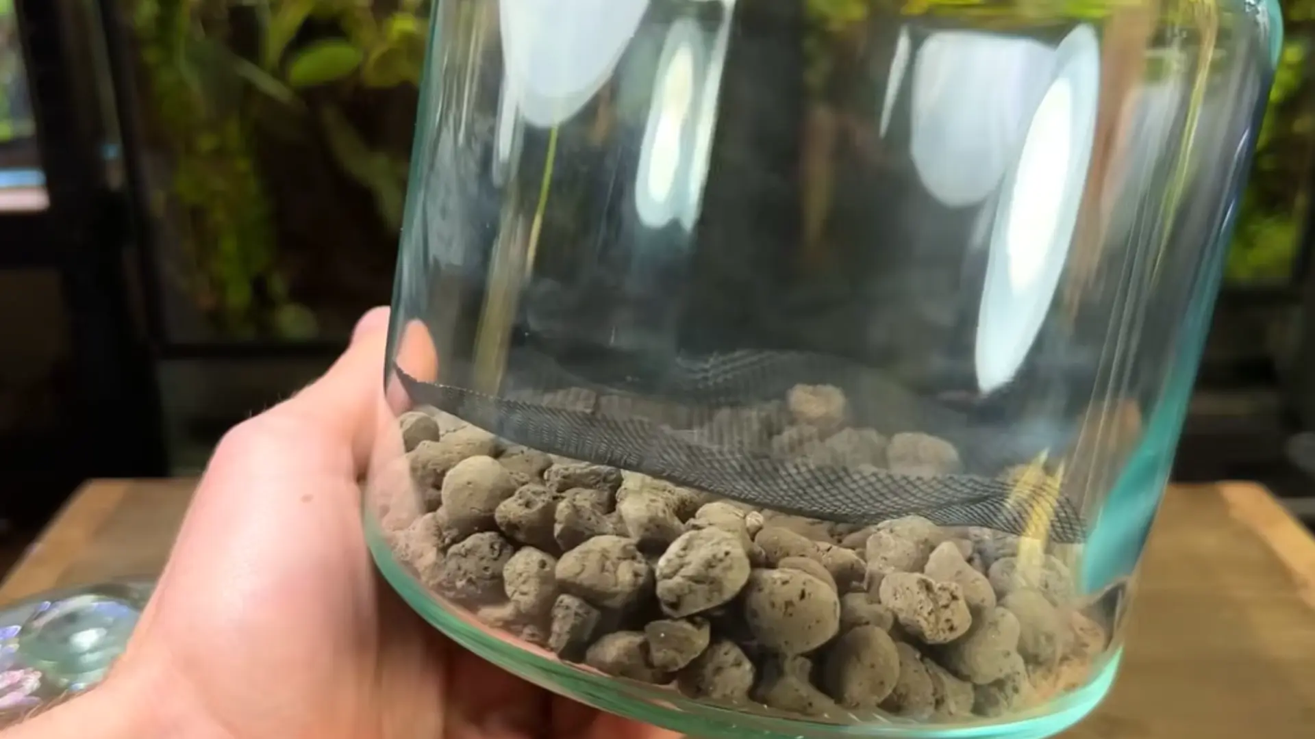 A hand holds a glass jar with rocks at the bottom and a mesh layer inside.