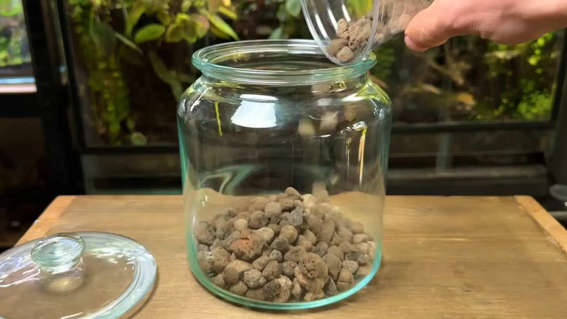 A hand pours small rocks from a jar into a glass jar, partially filled with rocks.