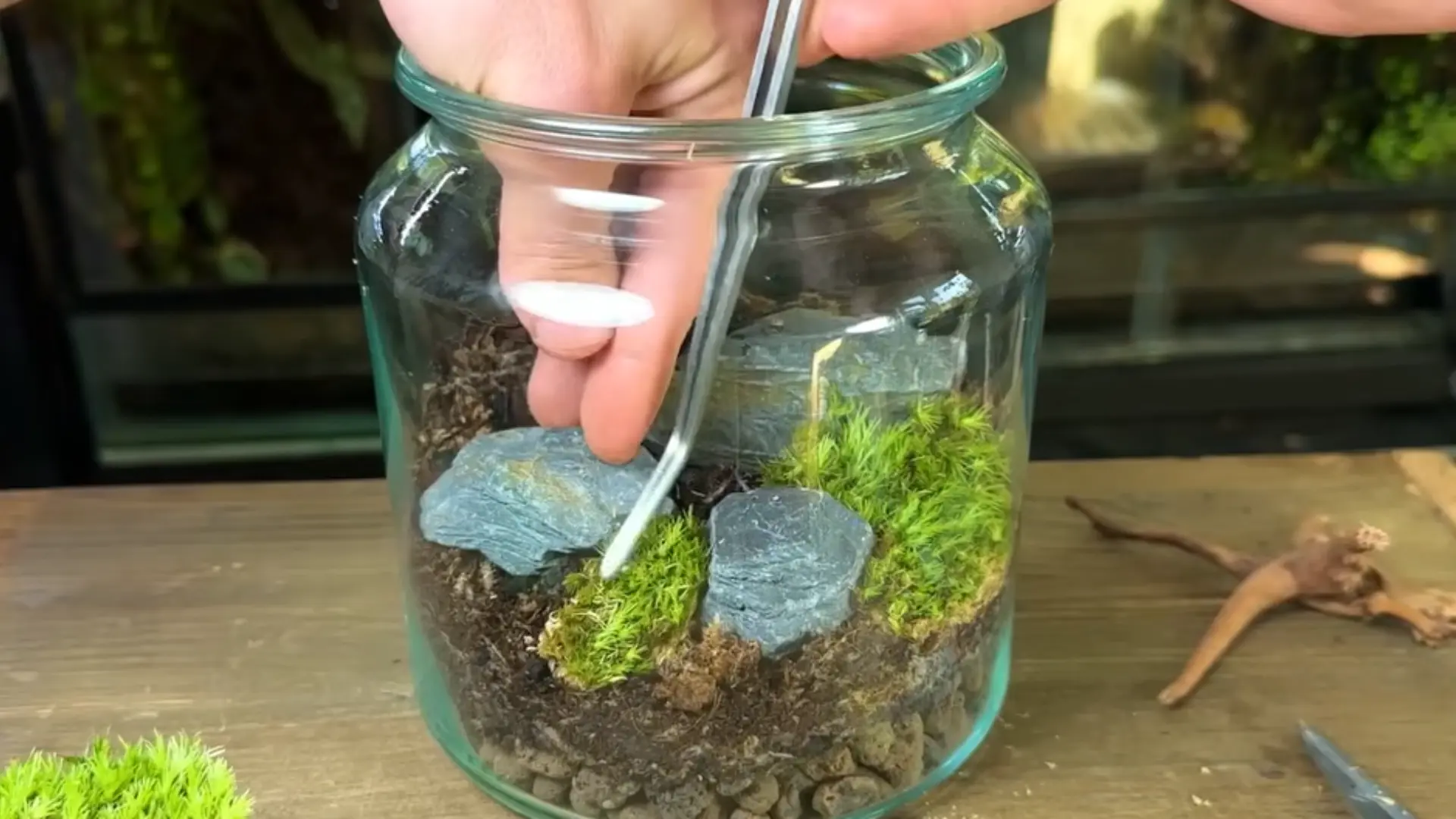 A hand uses tongs to place stones and moss inside a glass jar while creating a terrarium.