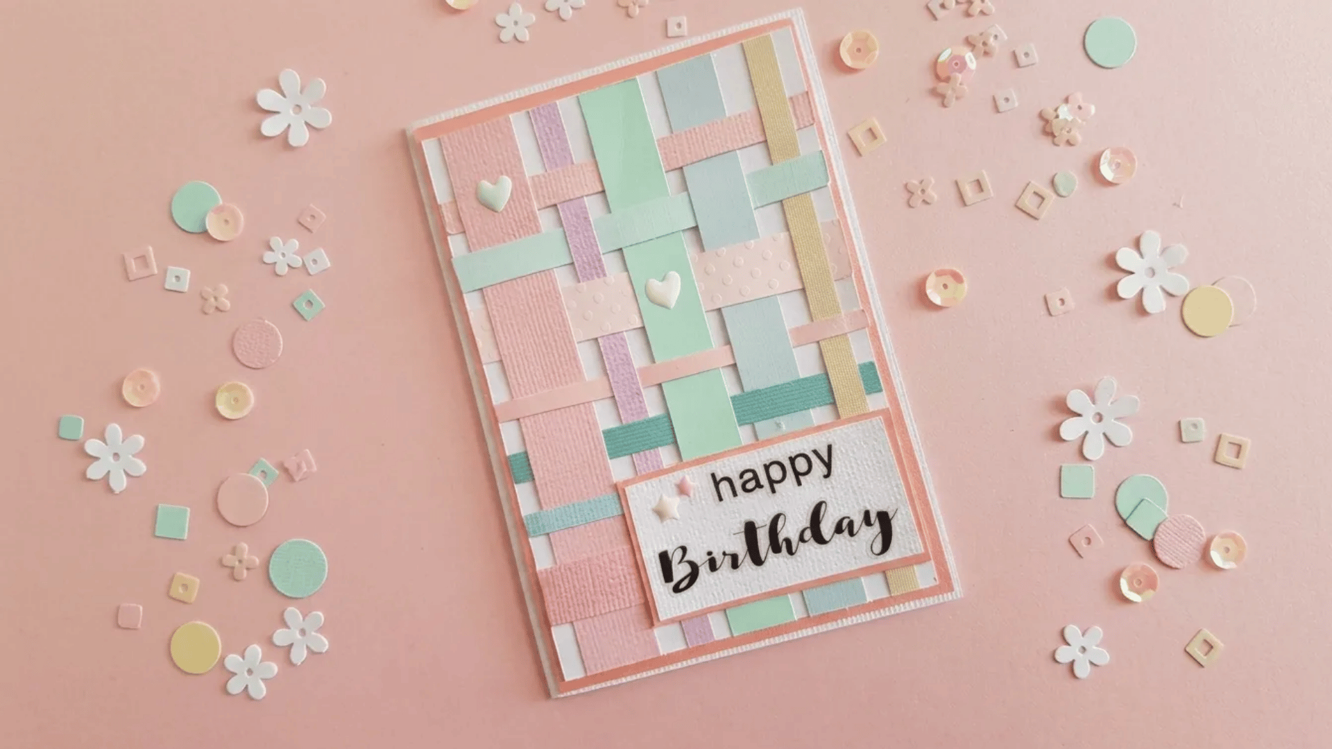 A pastel woven paper birthday card with layered strips, tiny heart accents, and scattered decorative sequins on a soft pink background.