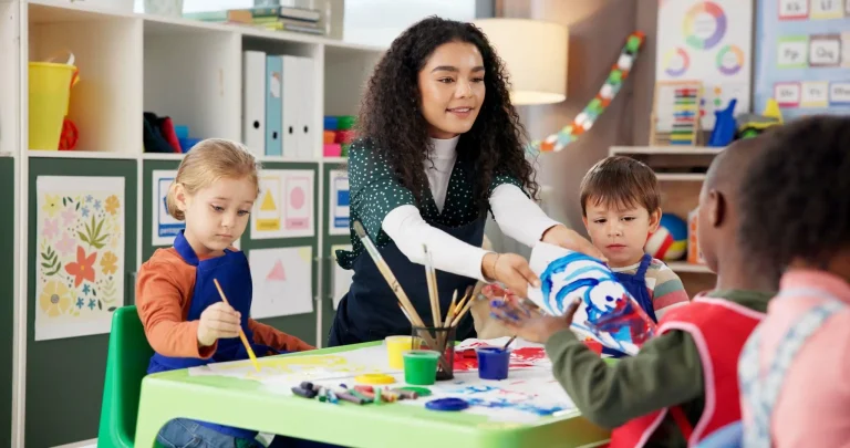 Teacher helping children with painting at a colorful table in a bright classroom setting