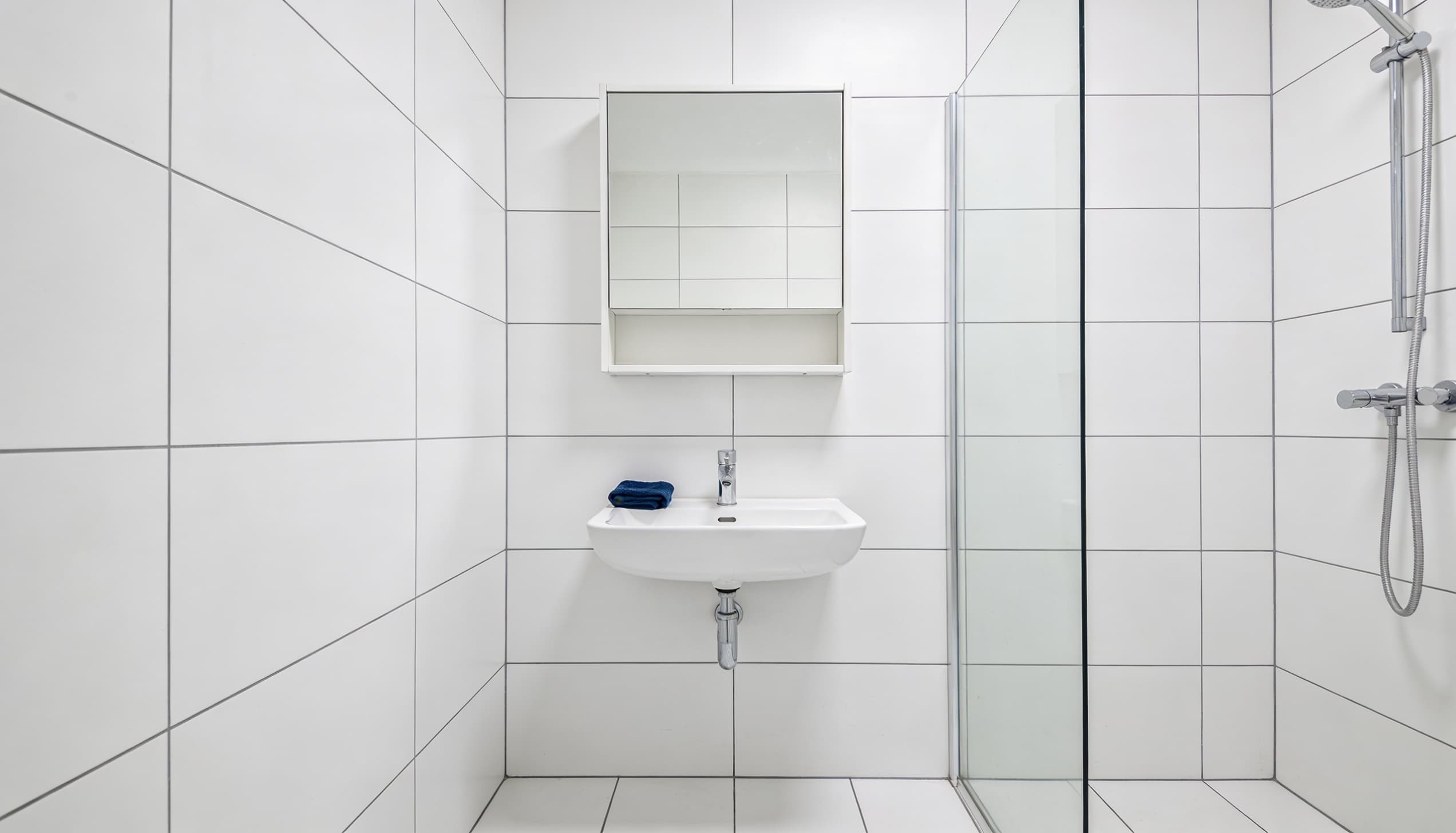 Minimalist Bauhaus bathroom featuring white square tile walls, a wall-mounted sink, a mirror cabinet, and a glass-enclosed shower.