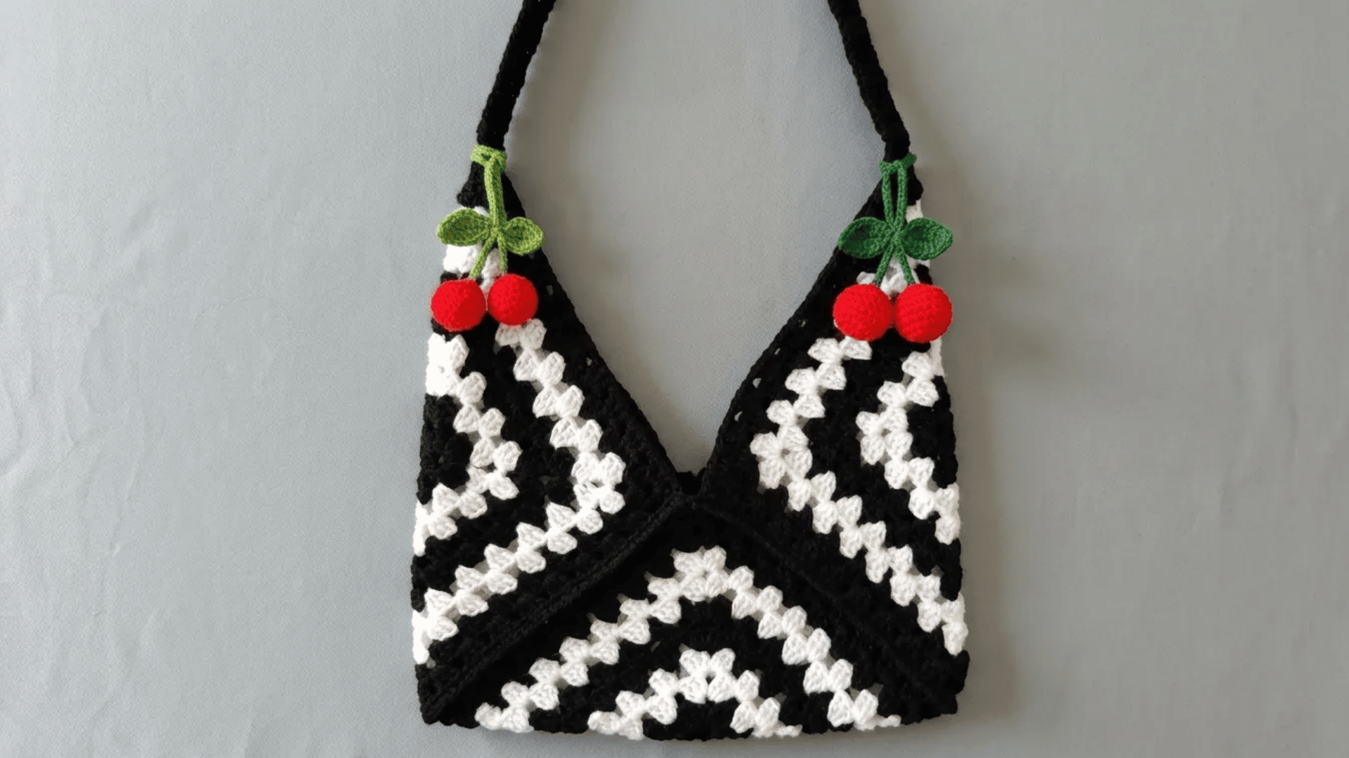 Black and white crochet tote bag with granny square patterns and cherry details hanging against a neutral wall.