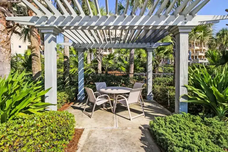 Outdoor patio set under wooden pergola surrounded by lush green plants and palm trees
