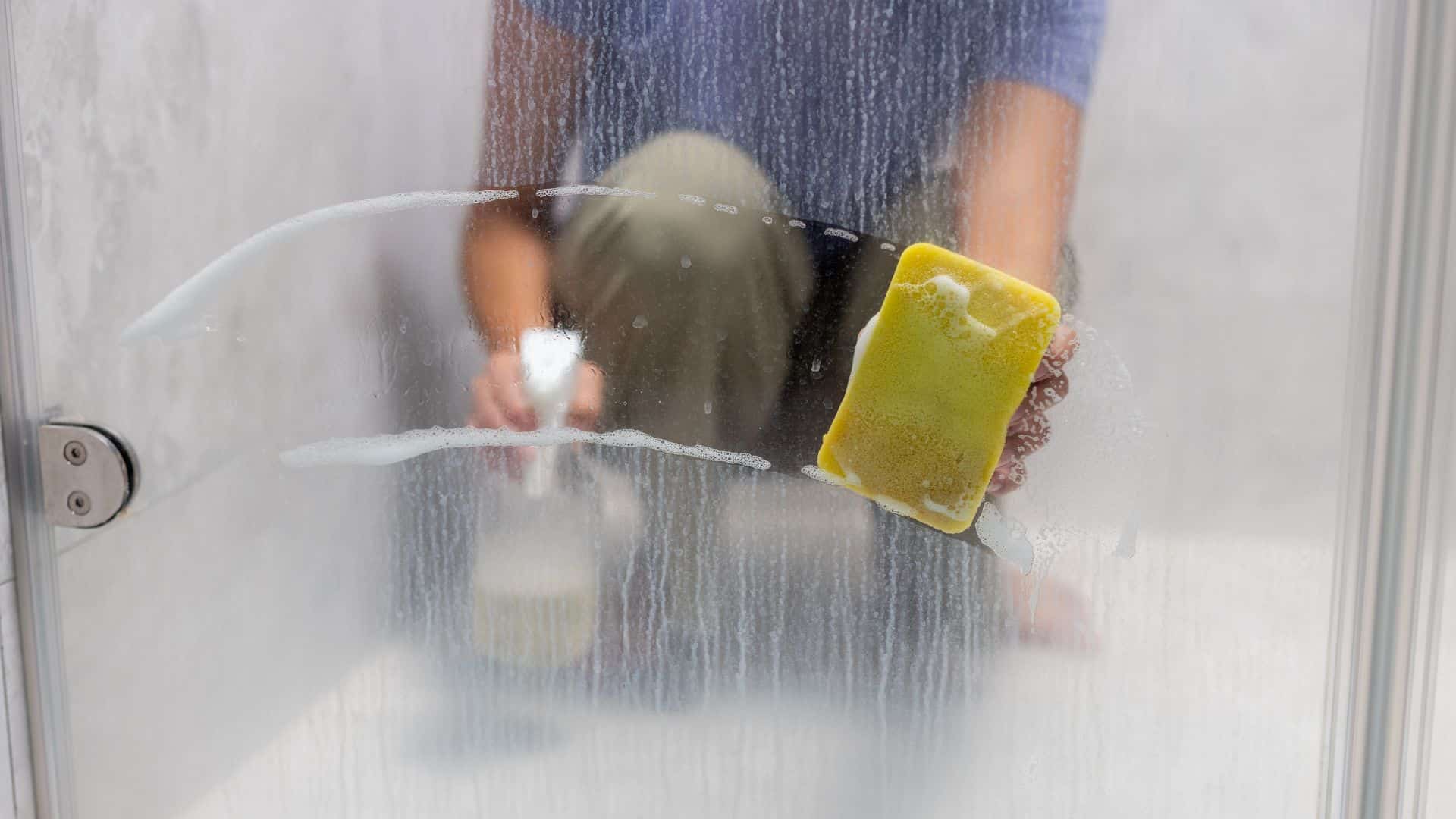 Cleaning shower glass with sponge and spray to remove hard water stains and soap buildup, leaving surface clear and streak-free