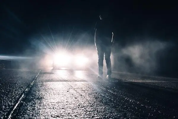 Silhouette of person walking towards bright car headlights on a foggy road at night