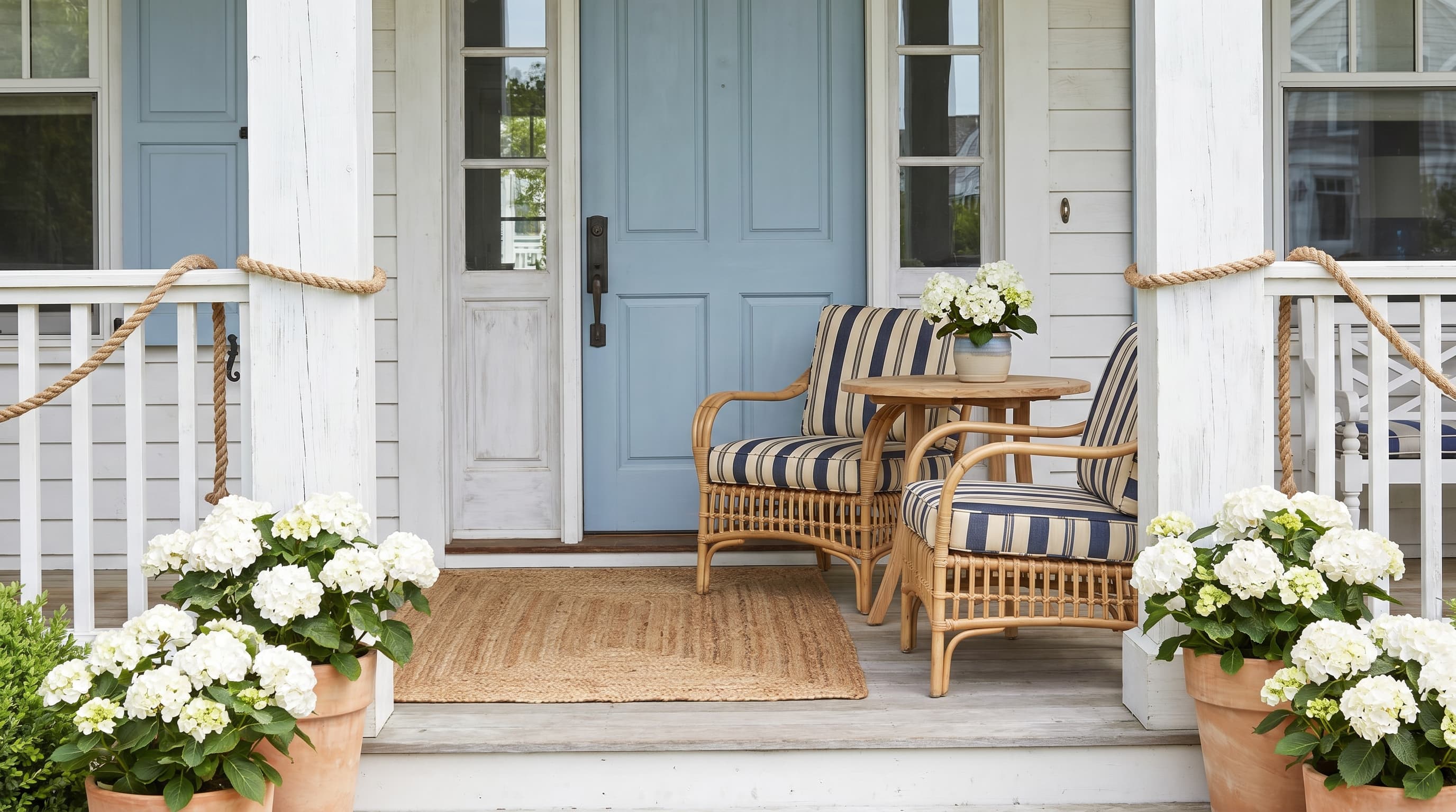 Coastal front porch ideas with soft blue door, rattan furniture, navy striped cushions, and potted white hydrangeas.