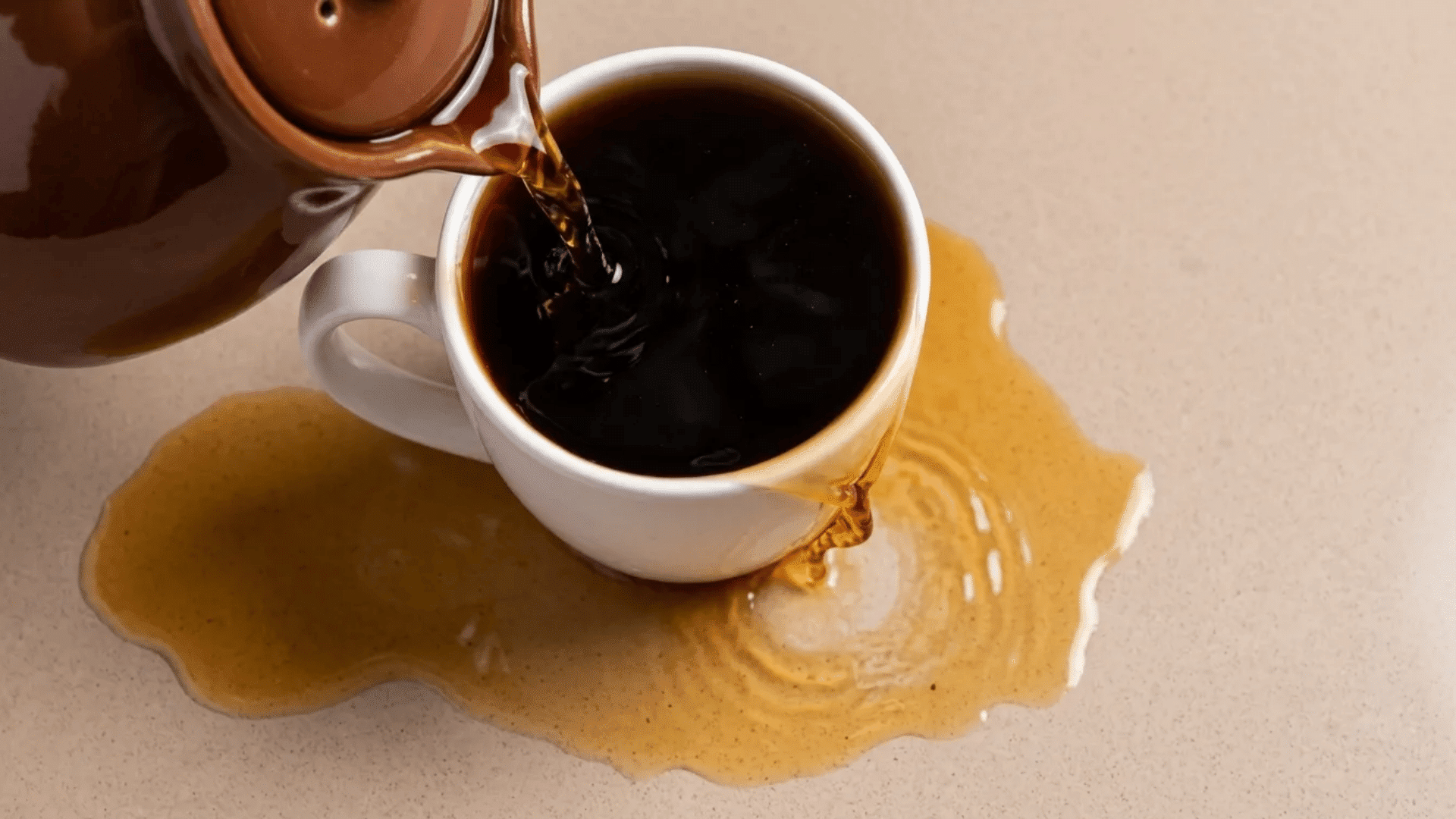 Coffee spilling from a white mug onto a light quartz countertop, creating a dark liquid stain and messy puddle across the surface.