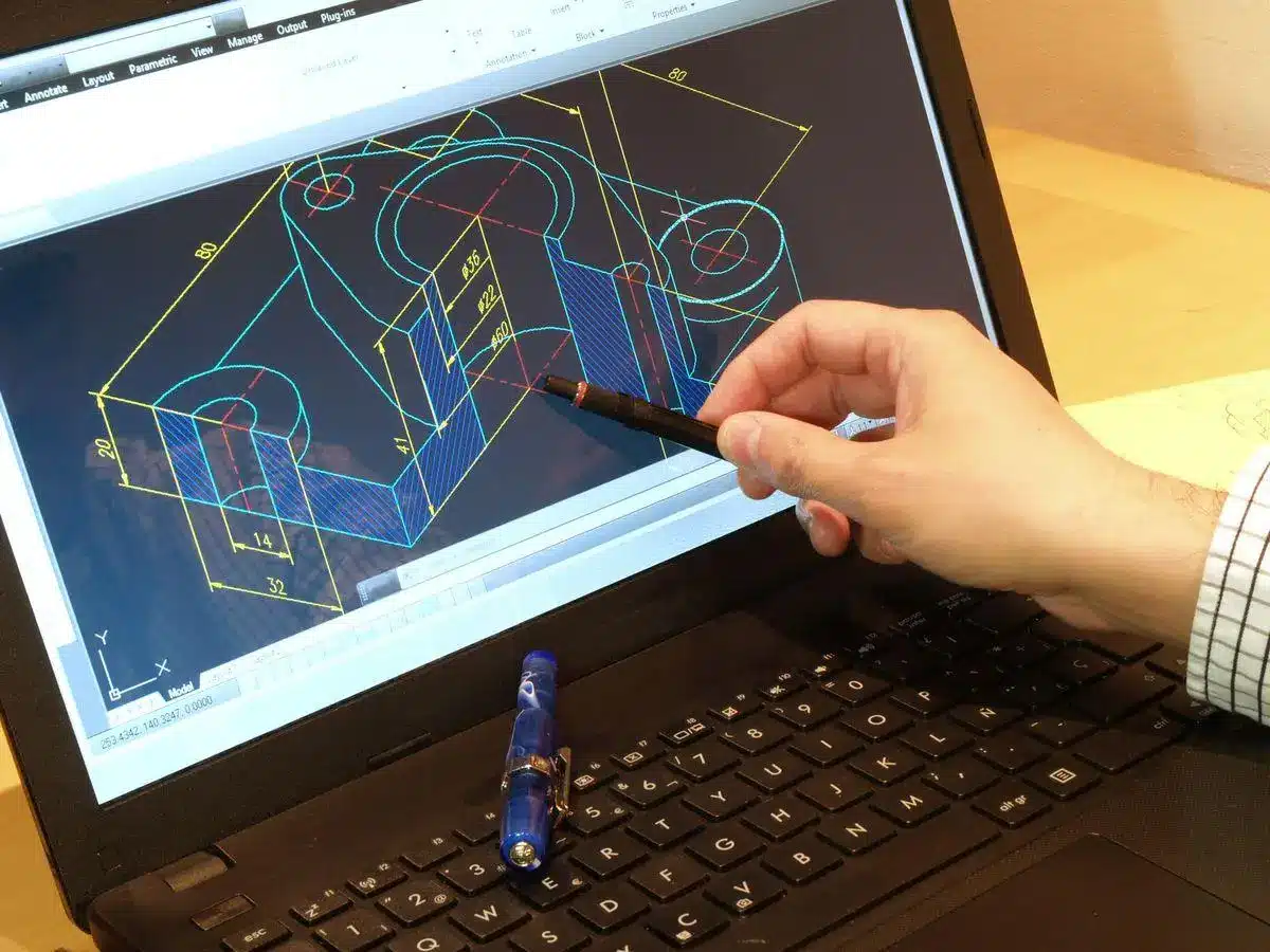 Hand pointing with pen at 3D CAD model on laptop screen in office setting