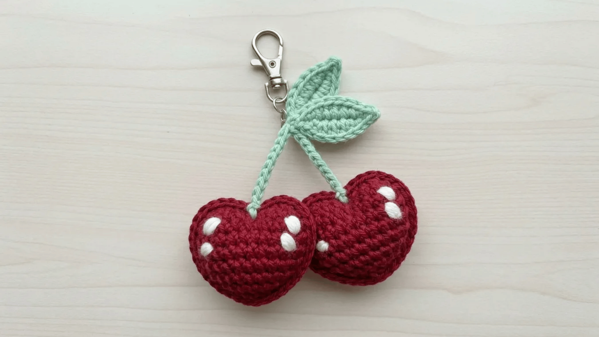 Cute handmade crochet cherry keychain with soft red cherries and mint green leaves on a light wooden surface.
