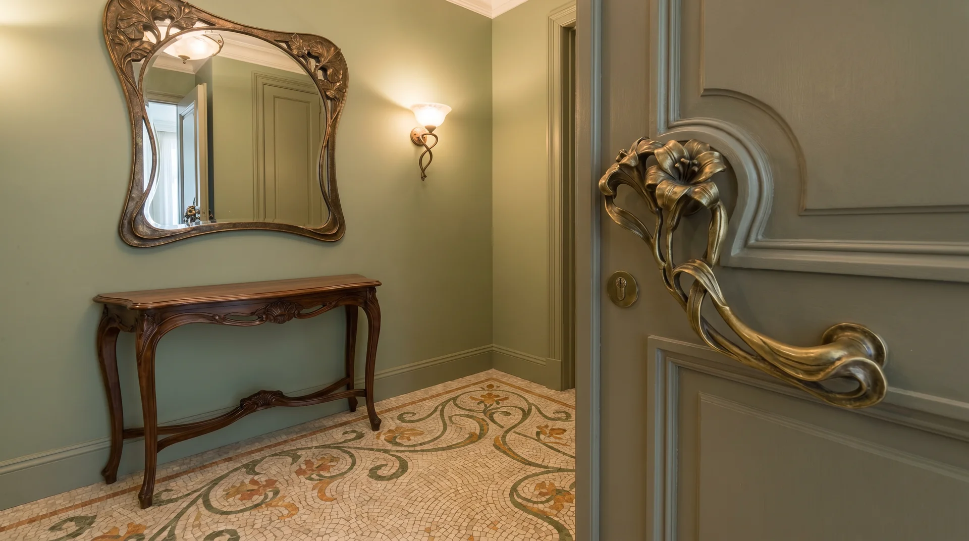 Art Nouveau entryway with an ornate gold door handle, a carved wood console table, a matching mirror, and tile floors.
