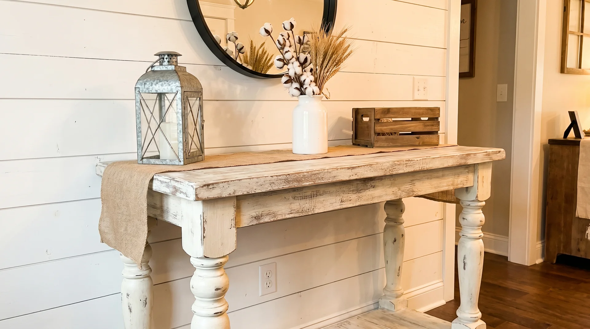 Distressed white farmhouse console table styled with a burlap runner, metal lantern, white vase, and wooden crate box.