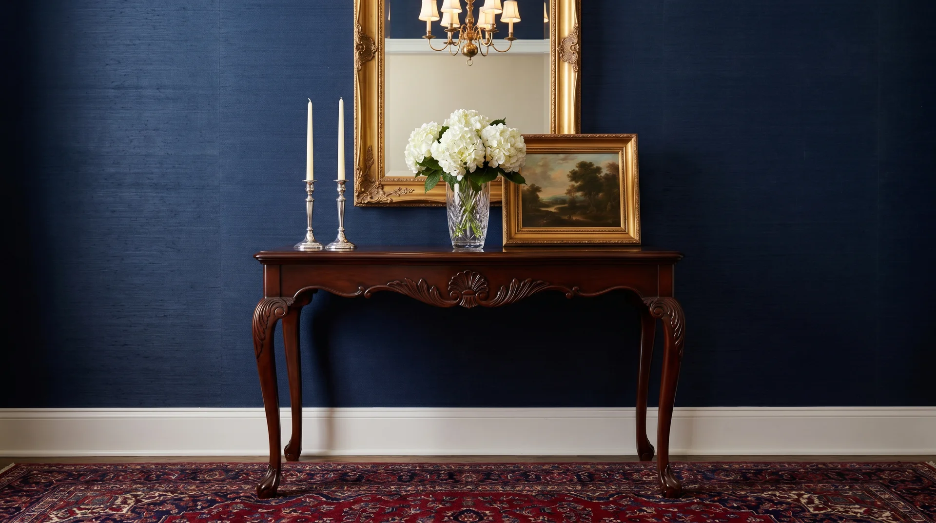 Traditional ornate wooden console table with a gold-framed mirror, white flowers, and tall silver candlesticks against a dark blue wall.