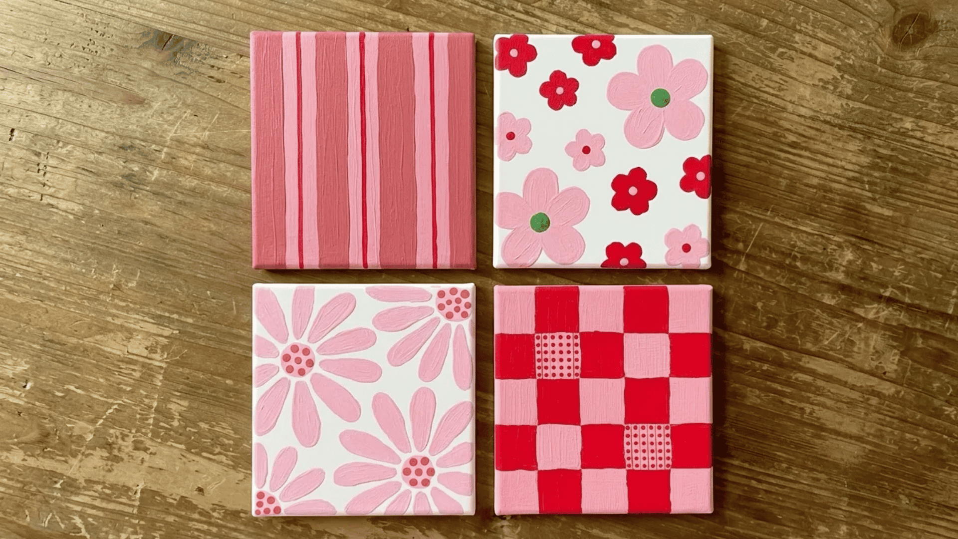 Four small pink-themed canvas paintings featuring stripes, flowers, daisy patterns, and a checkerboard design arranged on a wooden surface.