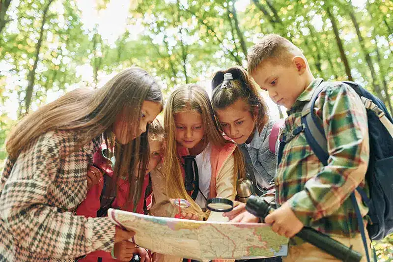 Children studying map in forest with backpacks and magnifying glass under sunlight