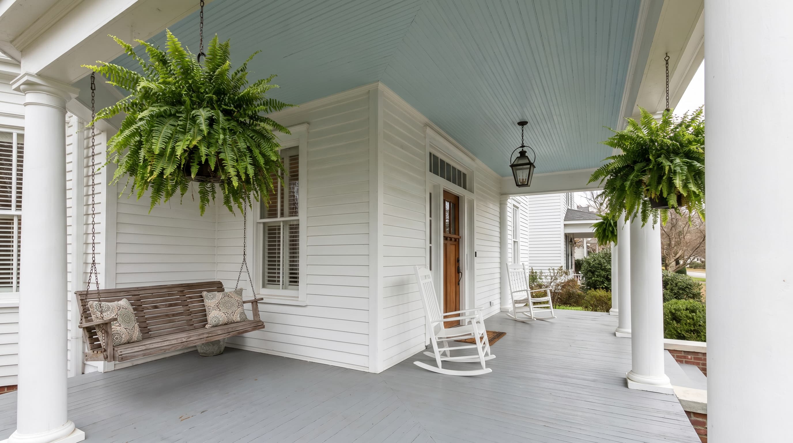 Front porch decorating ideas featuring a haint blue-painted ceiling, white railing, wooden swing, and hanging fern baskets.