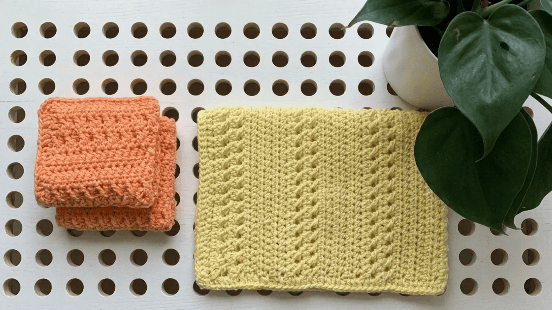 Handmade crochet dishcloths in soft peach and yellow tones placed on a white perforated table beside a green leafy plant.