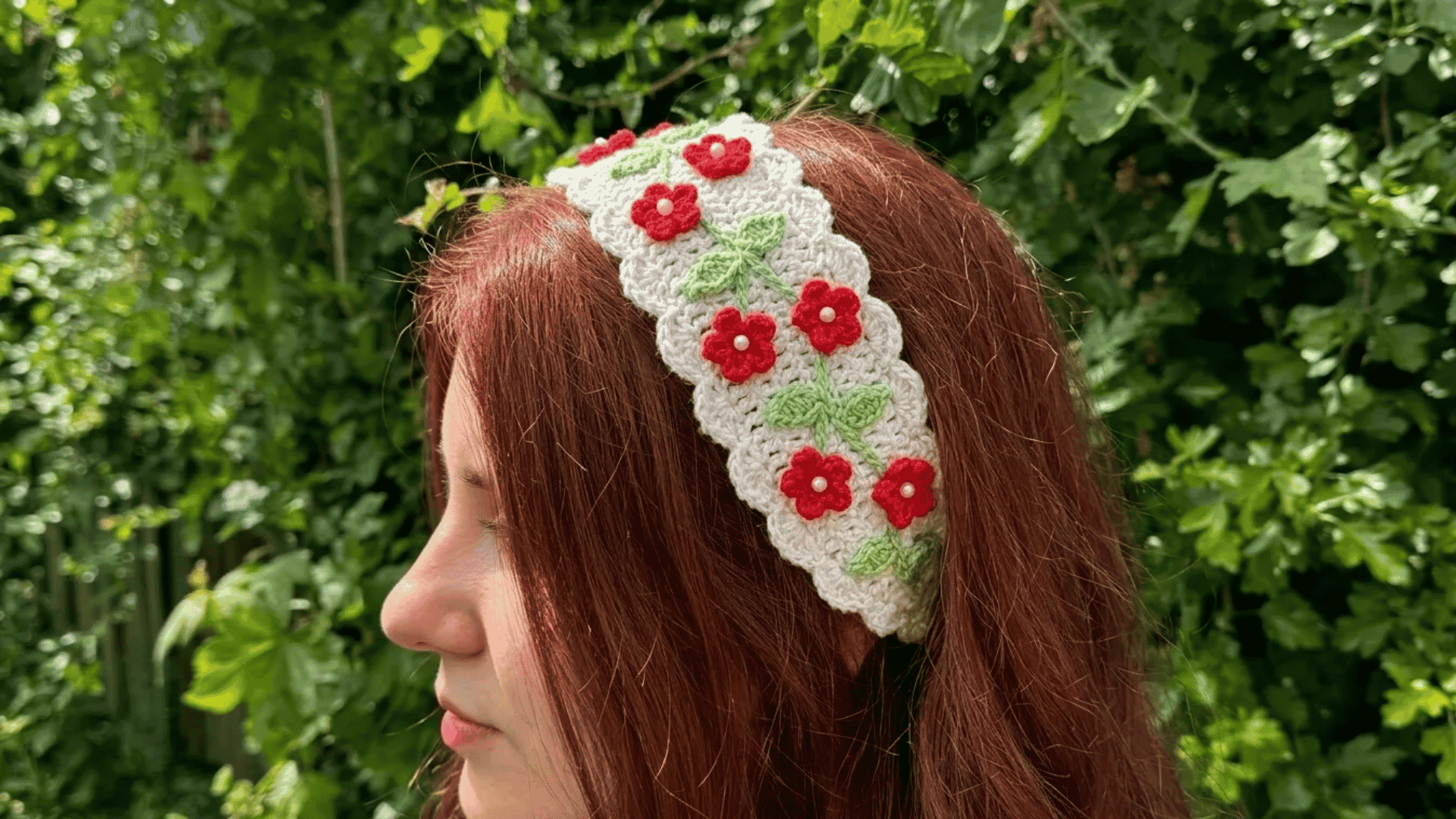 Handmade crochet floral headband with red flower details worn outdoors against a lush green background.