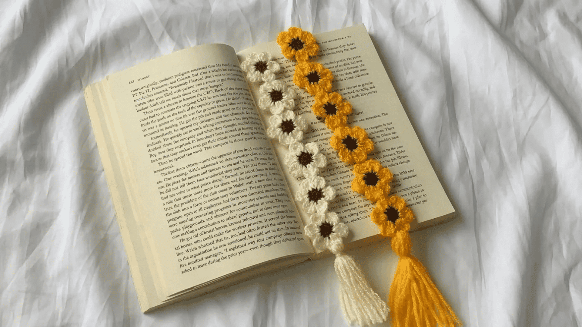 Handmade crochet flower bookmarks with tassels placed inside an open book on soft white fabric.