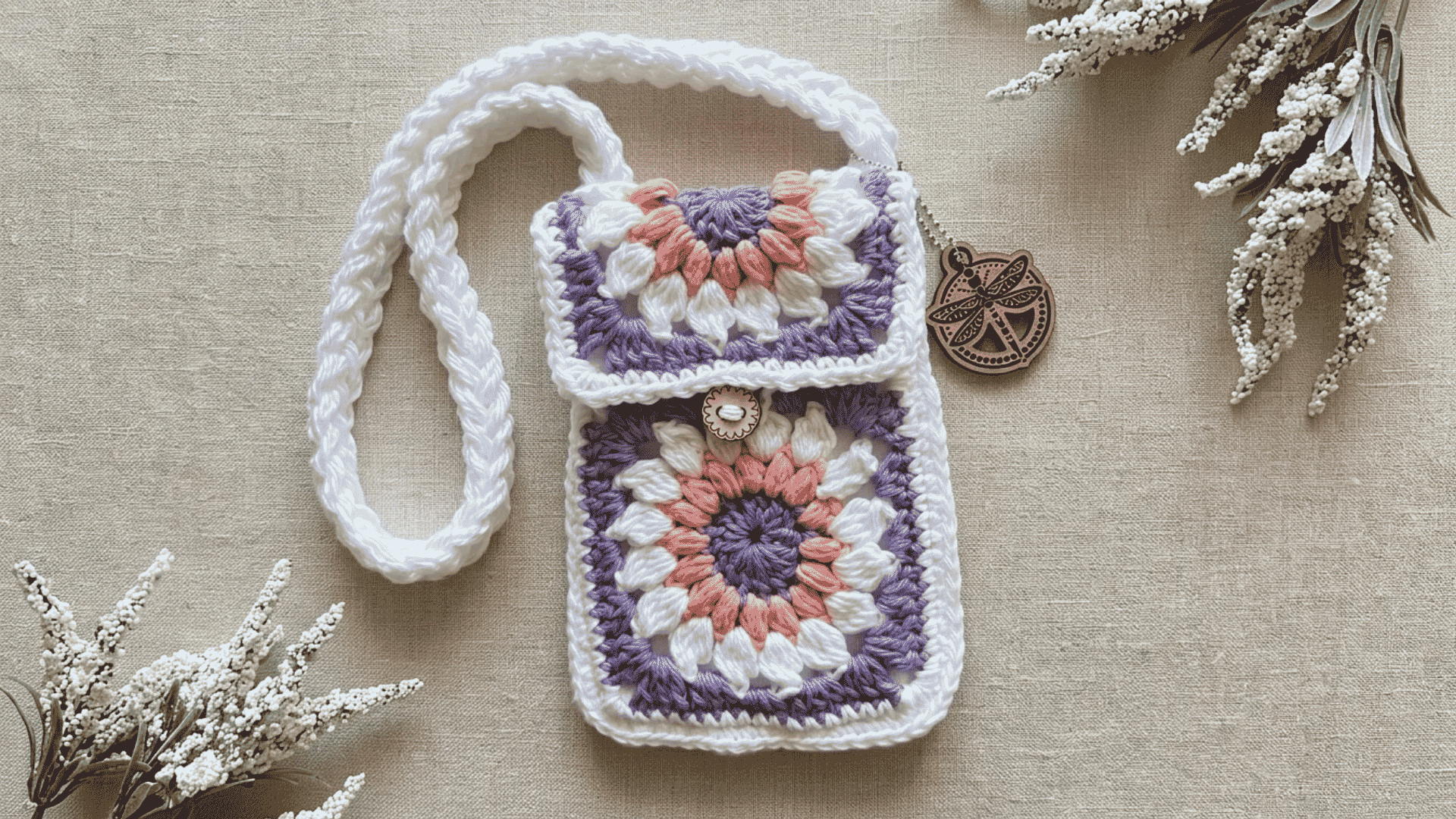 Handmade crochet phone pouch with pastel granny square patterns and a braided white strap on a neutral fabric background.
