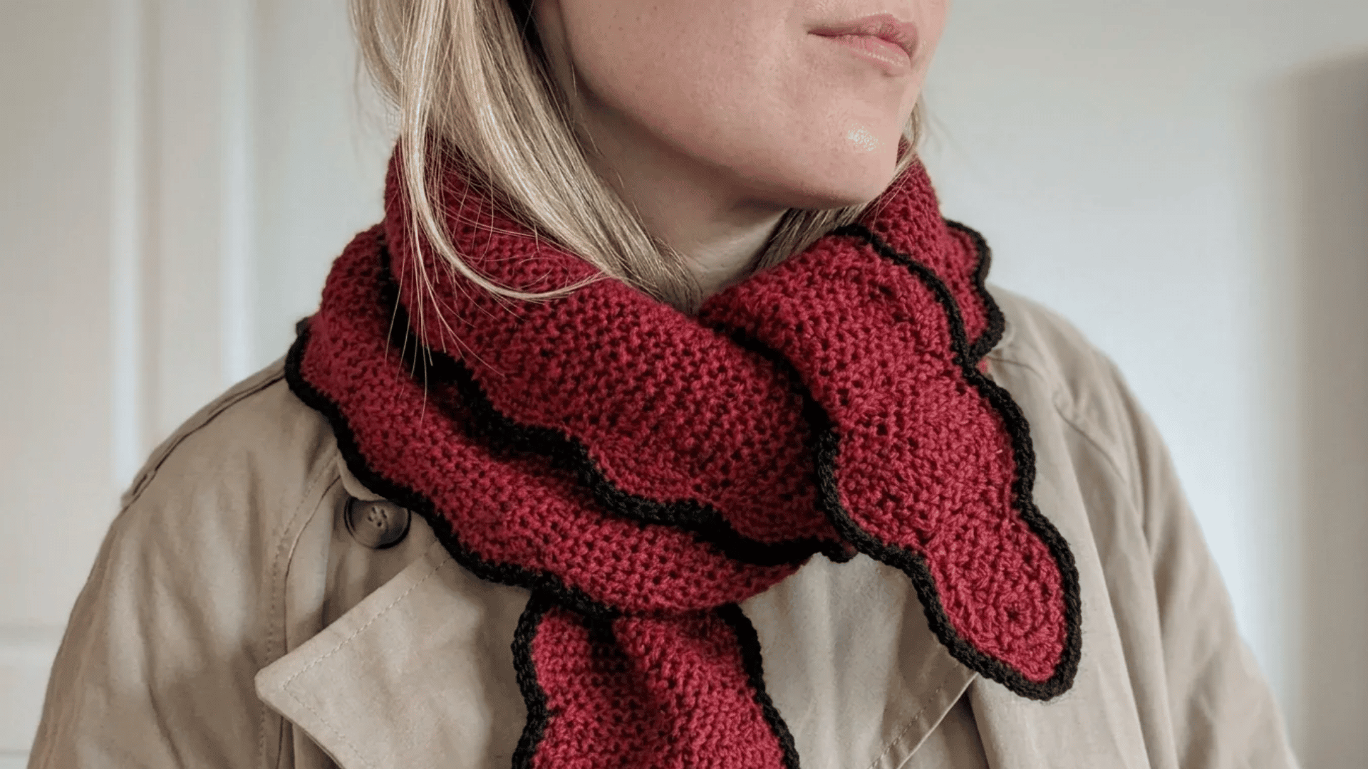 Handmade crochet scarf in deep red with black trim styled around a neutral trench coat.