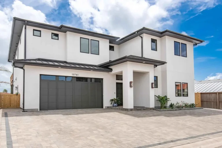 Modern two-story white house with dark garage doors and paver driveway under blue sky