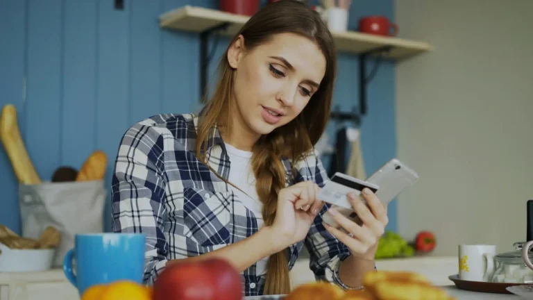 Woman in plaid shirt holding credit card and smartphone at kitchen table with fruits and coffee mug