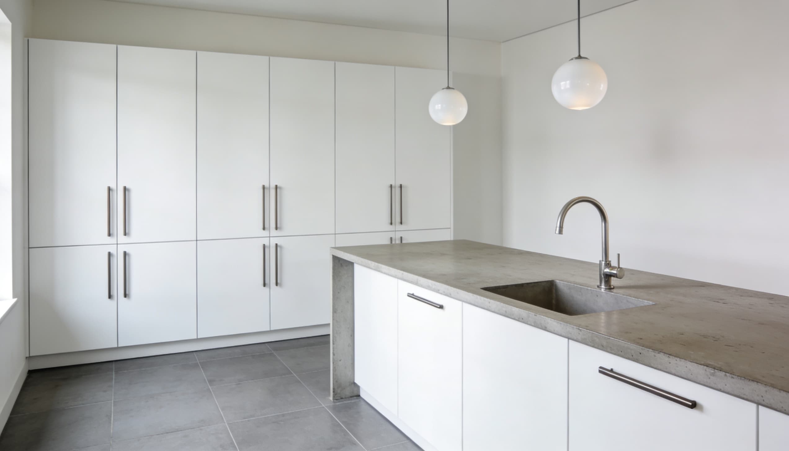 Bauhaus kitchen featuring white flat-front floor-to-ceiling cabinets with vertical bar handles, a light concrete island with a sink, and pendant lights.