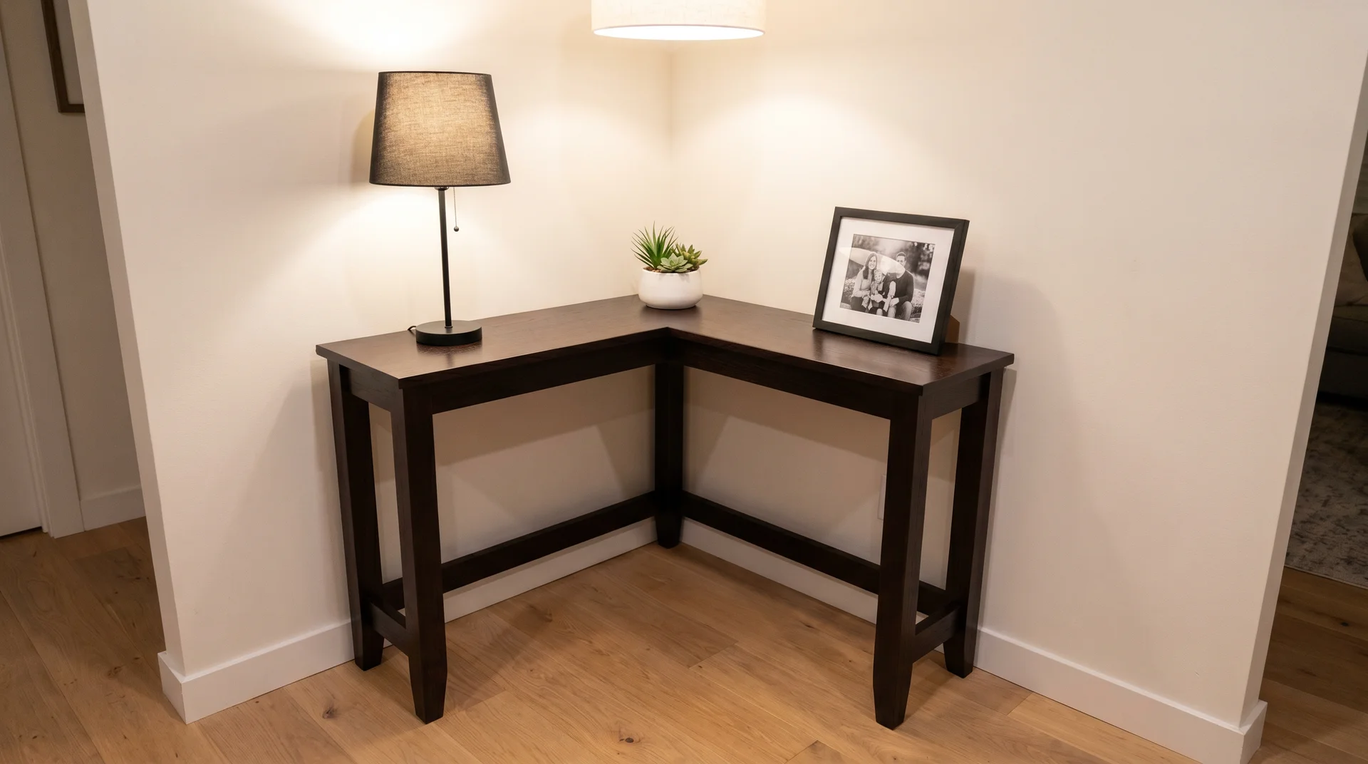 Dark wood L-shaped corner console table tucked into a wall corner, styled with a small lamp, potted plant, and a framed photo.