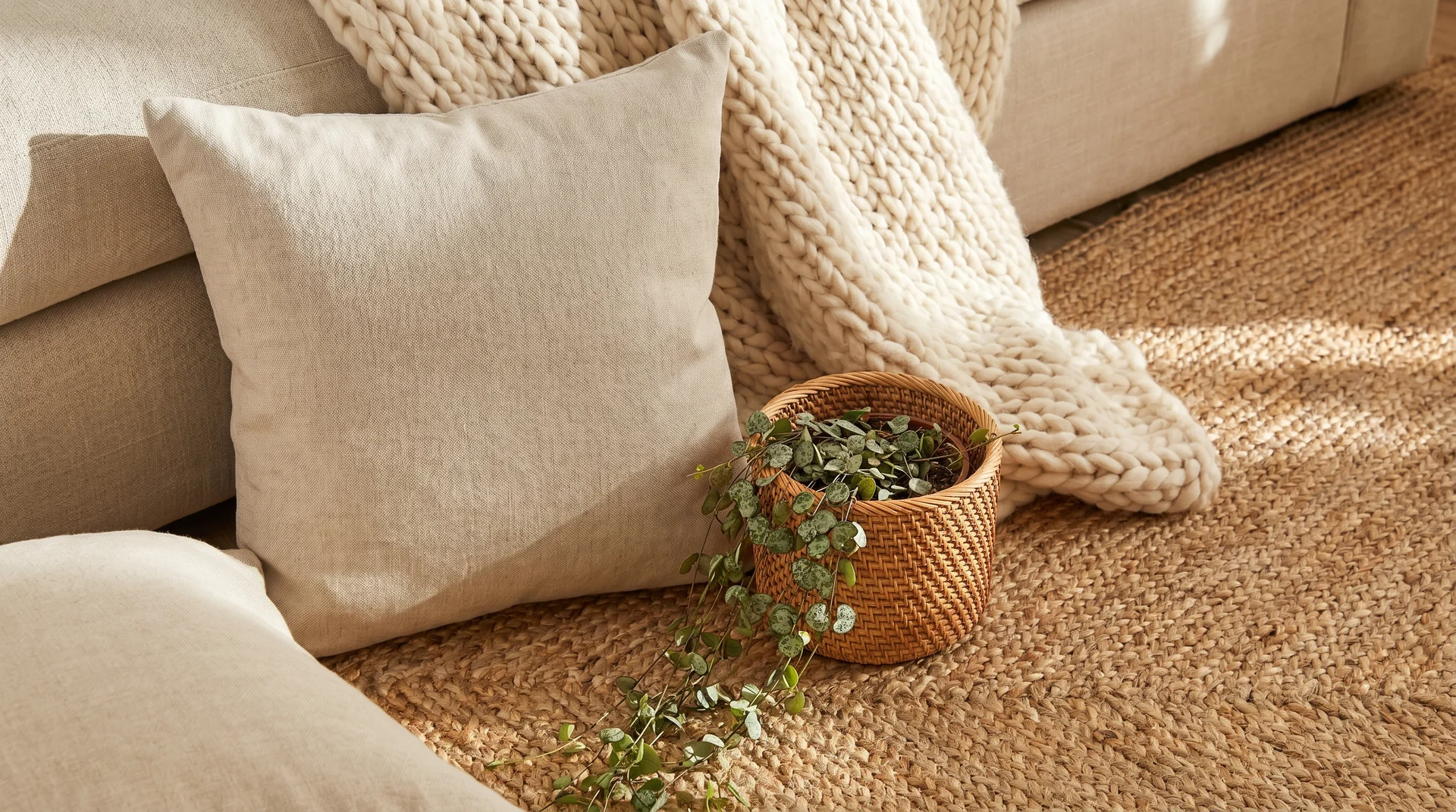 A close-up of a neutral sofa with a cream pillow, a chunky knit blanket, a woven rug, and a small rattan basket.