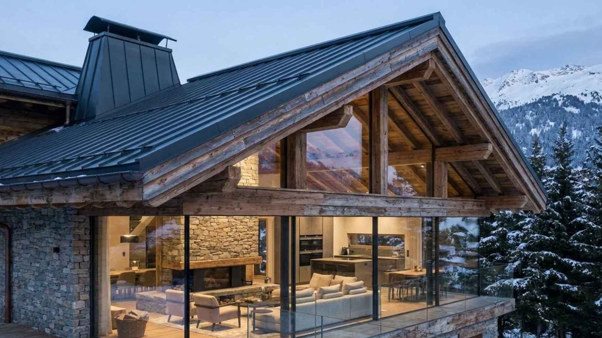 Modern alpine chalet with large glass windows, wooden beams, snow, and mountain scenery.