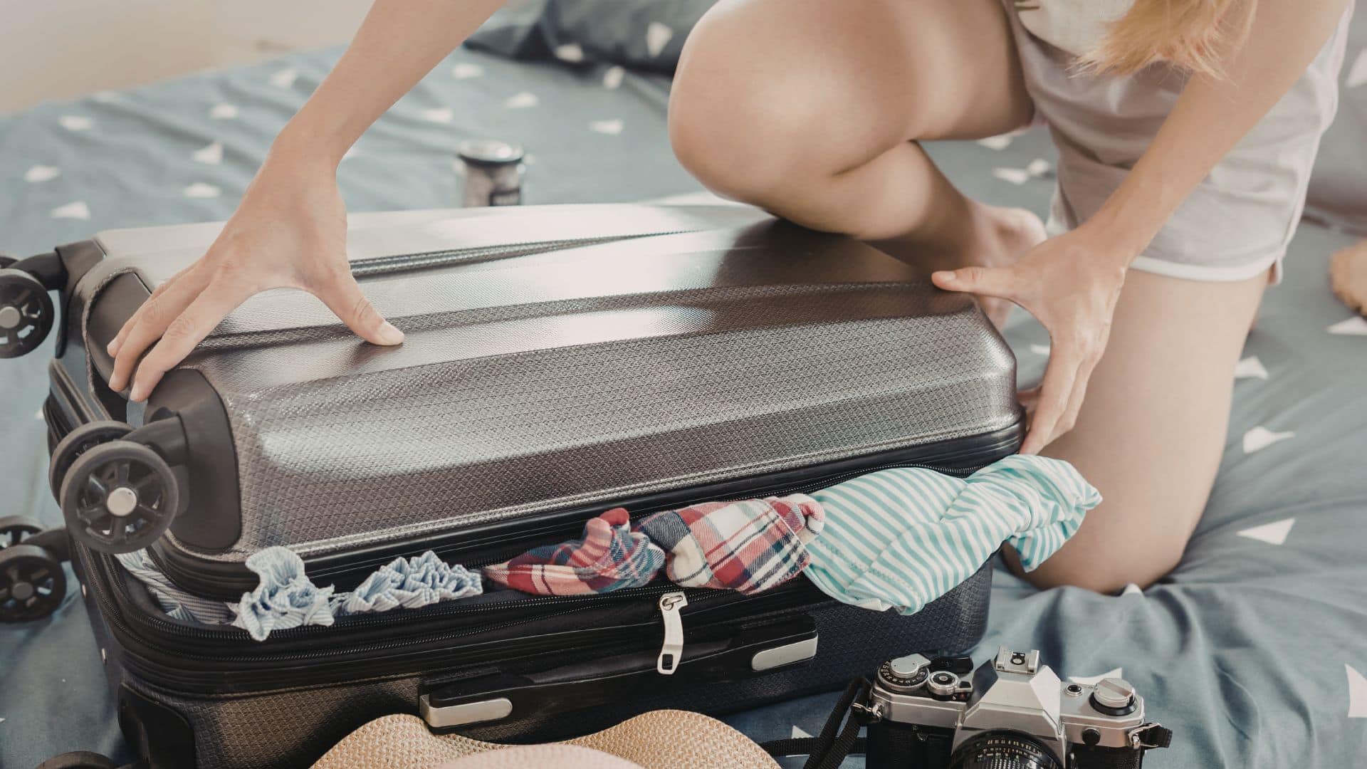Person struggling to close overpacked suitcase on bed, illustrating how to pack a suitcase efficiently and avoid overpacking mistakes