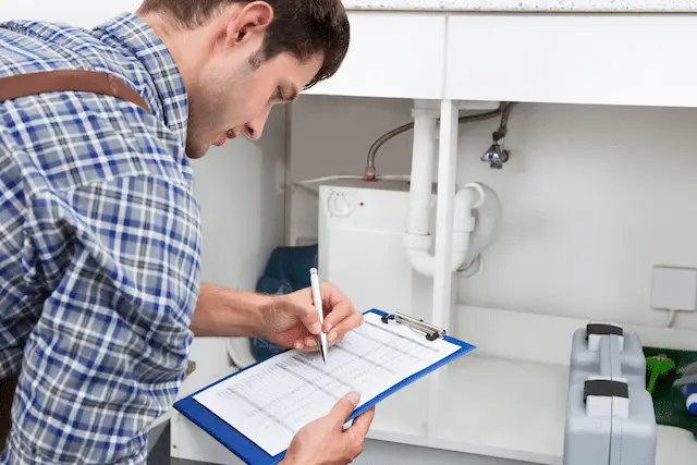 Plumber inspecting pipes and taking notes with clipboard under a kitchen sink