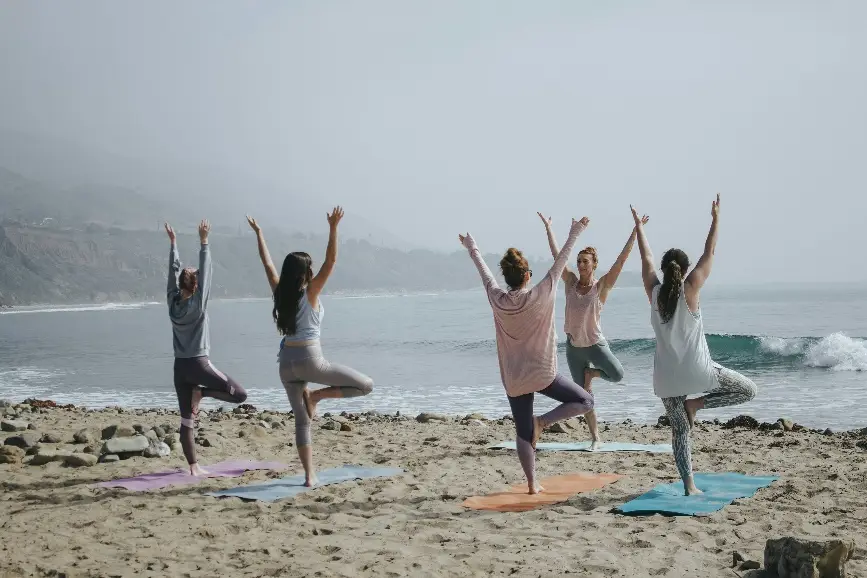 Five people practicing yoga poses on sandy beach by the ocean under cloudy sky