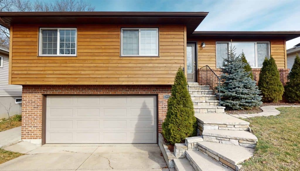 A split-level ranch house with light brown wood siding, a lower-level garage, and concrete steps leading to the entrance.