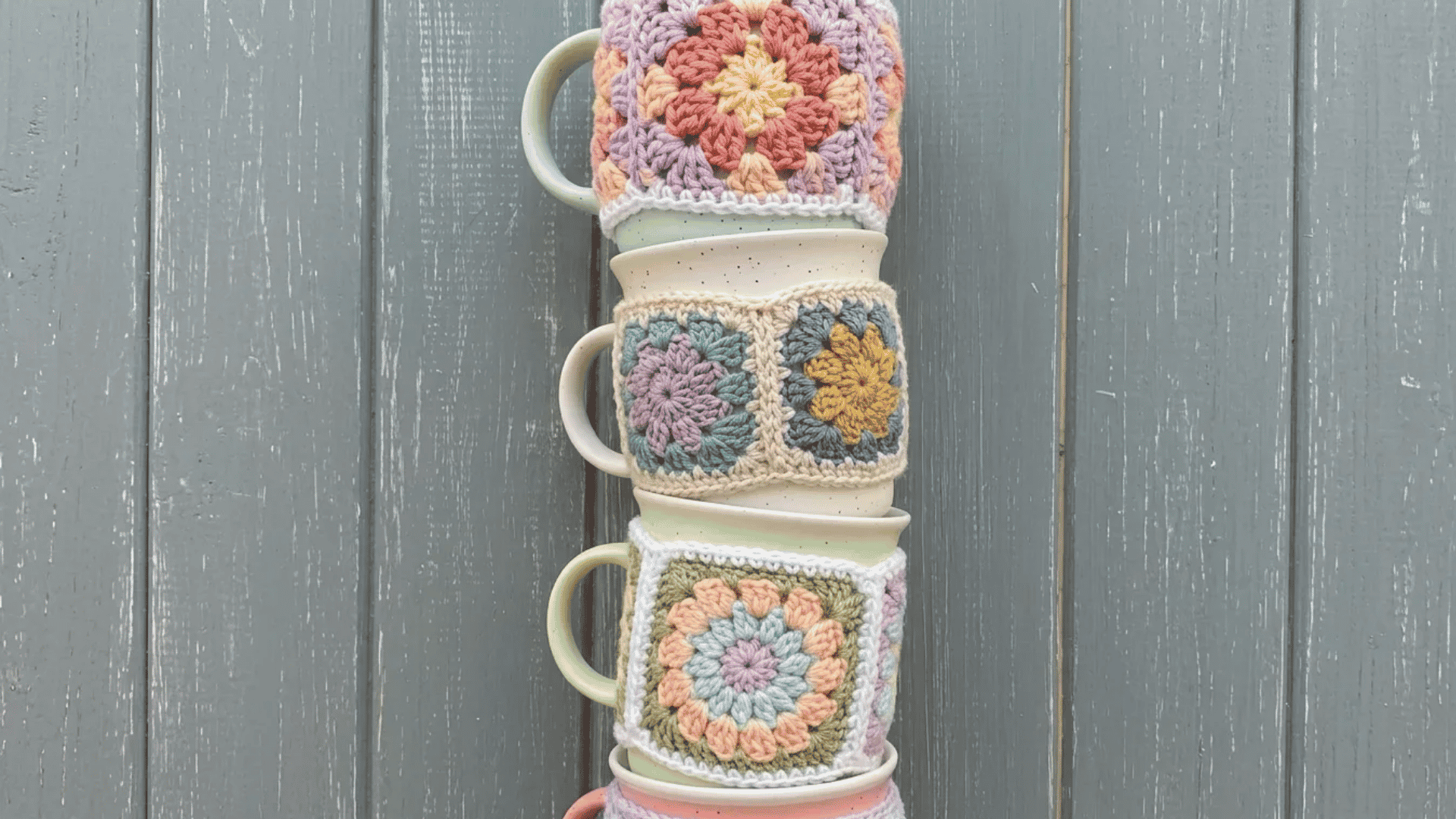 Stacked ceramic mugs wrapped in colorful granny square crochet cozies with soft pastel floral patterns.