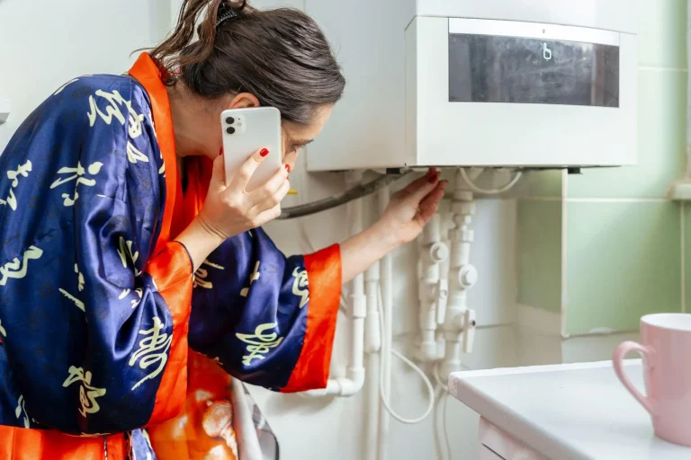 Woman in blue and orange robe inspecting wall pipes while on phone in a kitchen