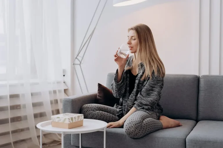 Woman sitting on gray sofa drinking water in softly lit living room